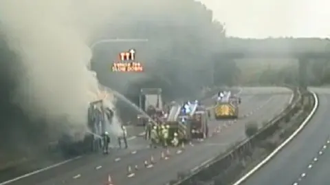 A blurry photo of a lorry stopped on the hard shoulder of an empty motorway. Smoke is billowing out the top and firefighters have hoses pointed at it. Three fire engines are stopped on the carriageway just ahead of it.