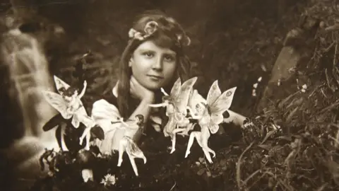 David Whittaker A black and white photo of a young girl with brown hair and a headband resting her hand on her neck and looking at the camera. In front are small dancing fairies. She is sitting outdoors with what appears to be a stream behind her.