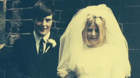 Supplied John on the left in a black suit and tie with a white flower on his jacket and Pat in a white wedding dress and veil