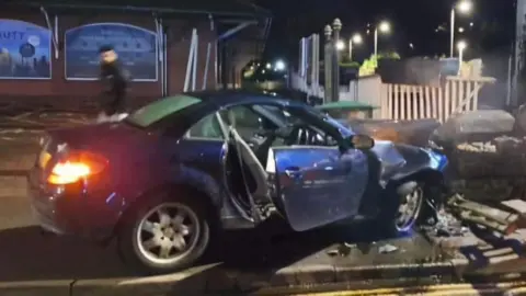 A blue car which has crashed into a low bridge above a stream. It is dark and the car's brake lights are on with the driver's door left open. The bonnet of the car is crumpled from the impact and there are bricks and glass shards across the pavement.