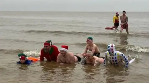 People in fancy dress taking part in a Christmas Day swim in Lowestoft. Six people are lying down in the water with two in the background standing up