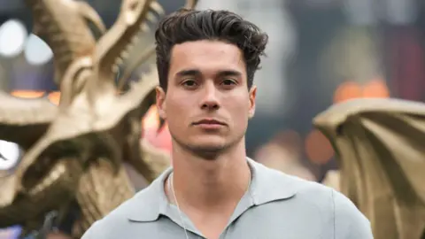 Miles Nazaire poses in front of a Dragon statue at a Premier. He has black hair up in a cwiff. He has a blue steel pose looking into the camera. He's wearing a chain and a grey polo shirt. 