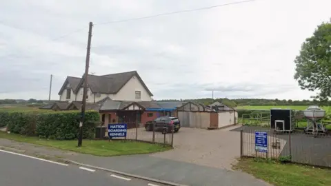 The image shows a building which appears to be a detached house and a driveway containing a black car and a trailer as well as a small boat on stilts. There are blue signs on a fence by the entrance to the driveway with white writing indicating the premises is Hatton Boarding Kennels.