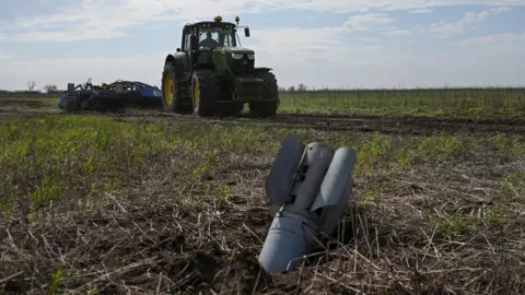 Reuters A light grey bomb with fins is seen protruding from the ground. In the background a large tractor is working on a farm field