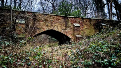 Jessica Eykel A large brick bridge, that has been repaired, showing bricks in place, and foliage all around it. 