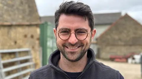 Janine Machin/BBC A man with dark hair brushed back. He has stubble and is wearing round spectacles and a black fleece. He is standing in front of farm buildings and is smiling at the camera.