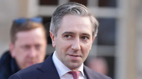 A close up shot of a man with grey hair, wearing a suit standing outside. Another man can be seen out of focus in the background.