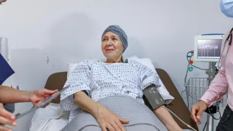 A stock image of a woman wearing a surgical gown and with a scarf over her head lying in a hospital bed with a blood pressure monitor on her left arm. Beside her is the screen the monitor is attached to and a man can be seen partly in the shot wearing a pink shirt and with a stethoscope around his neck. Only his arm and part of his chest can be seen.