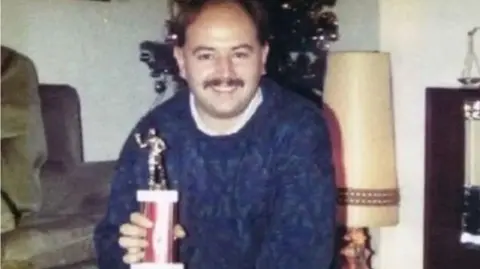 An old photograph of a man in a blue jumper smiling at the camera, he is holding a trophy with a footballer on the top