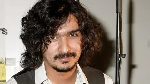 Getty Images Boyan Chowdhury, who left The Zutons in 2007, poses for a photograph wearing a waistcoat and white shirt. He has long, curly black hair and a goatee beard.