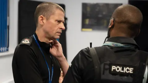 Northamptonshire Police Chief Constable Ivan Balhatchet, side on, talking to a police officer, who has his back to him. He has his hand to his neck, and has short dark hair. He has on a blue lanyard.