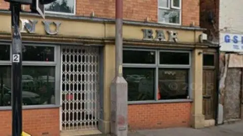 A bar sign which says Nu Bar above windows. The building has brown bricks and there is a grille over part of it. In front of the building are lampposts. 