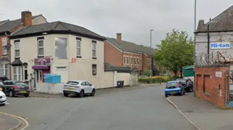 General view of Montgomery Street in Birmingham. Screenshot shows the junction of a road, on the corner there is a white two-storey building with a purple sign. On the right of the image there is a wall next to the rear view of a painted white brick building. On the side is a white sign with the words Hi-ton in blue
