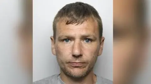 Darren Anderson looks at the camera in this police photo. He has short light brown hair and some facial stubble. He is wearing a grey top.