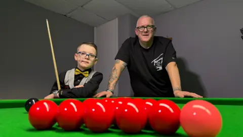 A young boy with glasses is wearing a black shirt, yellow bow ties and tweed waistcoat is holding a snooker cue and leaning on a snooker table, a man in a black shirt and tattoos on his arm is leaning next to him. In the foreground there are six red balls lined up. 