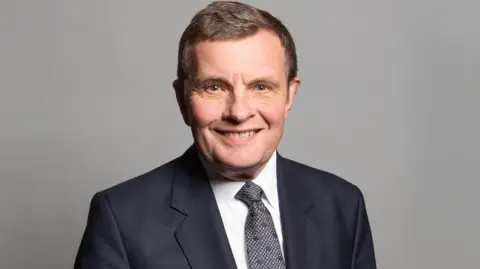 Profile photo of David Jones looking into camera wearing a dark suit, white shirt and grey tie, taken during his time in parliament.