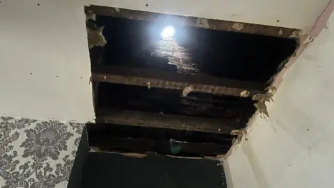 Kelisha Walker A large rectangular hole exposes wooden beams and brickwork above, with daylight shining through the roof. The surrounding ceiling shows rough edges and unfinished plasterwork.
