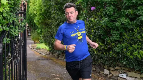 Getty Images Andy Burnham, wearing a blue t-shirt with a bee on it, jogging out of a black gate.