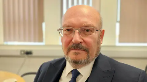 Paul Moseley/BBC Graham Plant is looking at the camera. He's wearing rimless glasses, a white shirt, blue tie and navy/grey jacket.