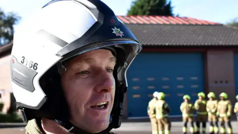 Simon Carr Retired firefighter Simon Carr in Nottinghamshire Fire Service helmet in front of colleagues at a fire station.