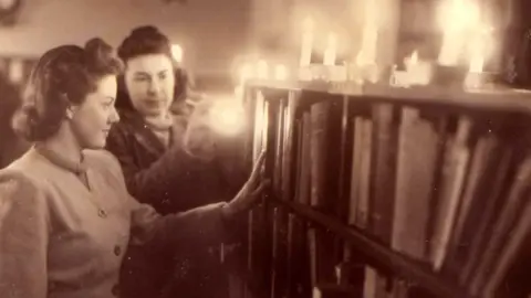 A black and white image of two women looking at a bookcase. There are candles on top of the bookcase. 