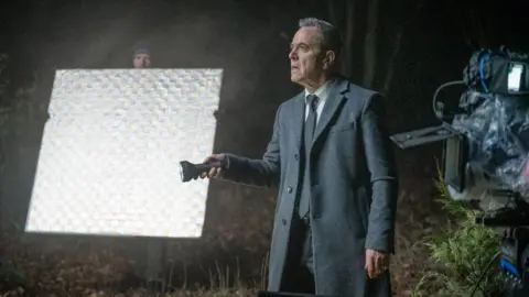 Run Away Simon Greene, played by James Nesbitt, is holding a torch and staring off camera in this candid image during filming in the woods. There is filming equipment on each side of him. 