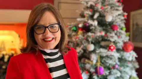 Lewis Adams/BBC Jane Malyon smiling at the camera while standing in front of a Christmas tree in her living room. She is wearing a black and white striped jumper with a red jacket over the top.