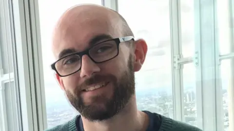 A man wearing a green jumper and glasses and smiling. Behind him is the London skyline, seen through the glass of an elevator.