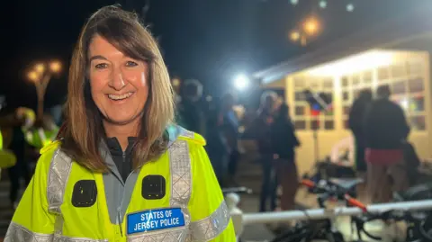 Alison is wearing a bright yellow reflective jacket with a patch that reads “STATES OF JERSEY POLICE” stands outdoors at night. Behind them, there are bicycles, a lit building, and several people gathered under string lights.