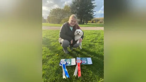 Melissa Taylor A woman wearing a coat crouching on the grass in front of two medals and certificate. She is also holding her dog King Charles Spaniel.