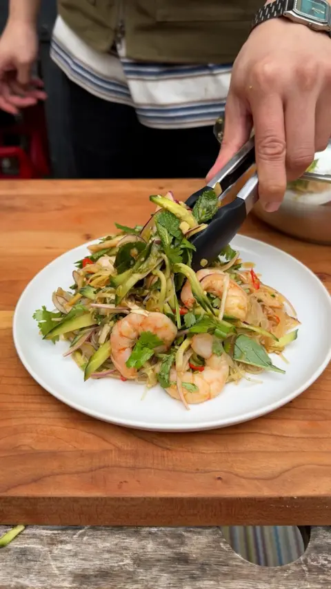 A hand holding a pair of tongs reaches down to serve portions of a green salad with noodles and prawns.