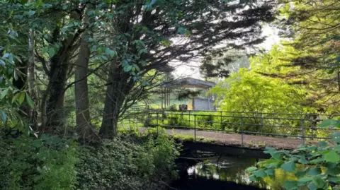 Part of the area, with a bridge over a river, trees, sun shining through leaves and part of an old barn in the background.