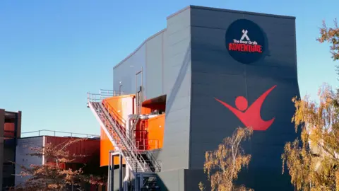 A large grey building has a sign that reads The Bear Grylls Adventure above a red logo of a person jumping. On the side of the building is a long metal staircase.