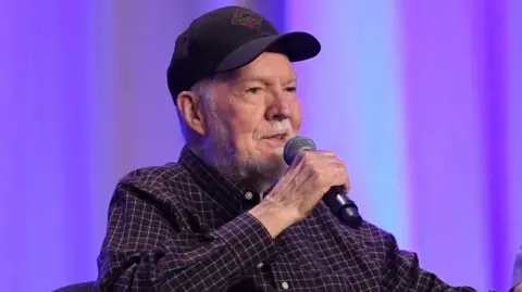 Getty Images An image of Peter Renaday sitting in front of a purple curtain wearing a checked shirt and a cap, at an event in 2017