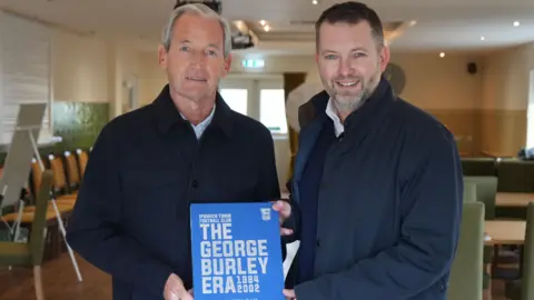 Jamie Niblock/BBC George Burley y Neil Prentice se paran al lado de un libro azul llamado 