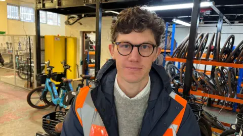 A man in a hiviz vest is looking at the camera infront of some ebikes