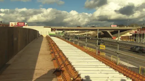 A motorway bridge with traffic flowing, with a long metal structure sitting alongside the motorway in the foreground