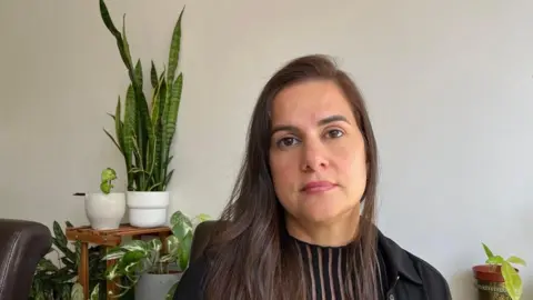 A head and shoulders image of Ana who has long, dark hair and is wearing a black top underneath a black jacket. She is sitting in a living room and there are various house plants on shelves in the background. She has a serious expression on her face.
