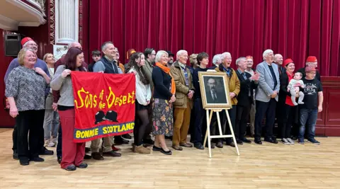 Amanda Dewar James Finlayson's family and fans gather around a new framed portrait of the actor in front of a stage with red curtains. The oil painting is mounted  on an artist's easel. Two people are holding up a banner showing Laurel and Hardy's famous bowler hats and the words Sons of the Desert, Bonnie Scotland. 