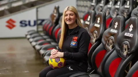 Fixed Point Media A woman with long, straight blonde hair and wearing a black tracksuit sitting in a dug-out at the MK Dons ground. She holding a yellow football on her knee. Empty red-and-grey seats are around her.