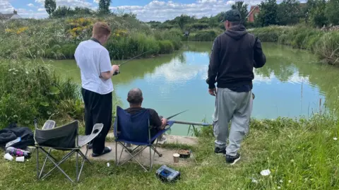 Recovery Rods Three men fishing together at a lake, with their backs to the camera as they look out across the water. Two are standing and the one in the middle is sitting in a camping chair