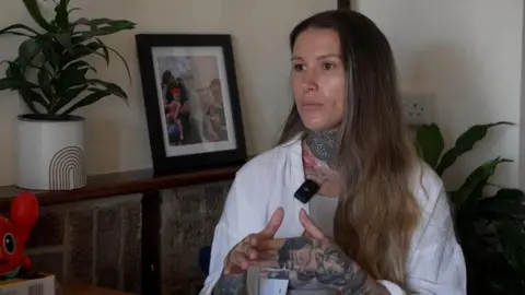 Carly sitting at a table being interviewed for television. She is wearing a white jacket with a microphone attached and has several prominent tattoos.