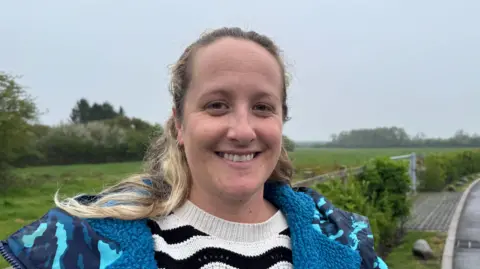 A woman smiles for the camera with green fields and a metal gate behind her. Sam Whicker is wearing a black and white hooped jumper, with a multicoloured winter coat over the top. She has brown and blond long hair which sits on her shoulders.