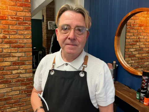 BBC News A man with blonde hair and wearing glasses is standing in front of a mirror. He wears a black apron with hair clips attached to one side. 