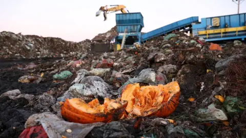 Reuters A dumpit site filled with food waste. A broken pumpkin is in the foreground. In the background, there is a yellow excavator with a grapple attachment above a blue skip leading to a conveyor belt.