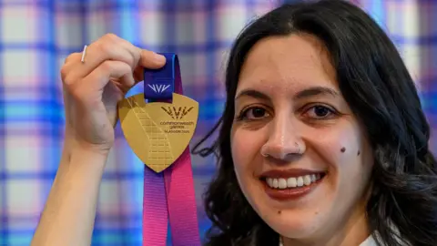 Militsa Milenkova - a woman with dark hair down to her shoulders, smiles while holding up a gold medal.