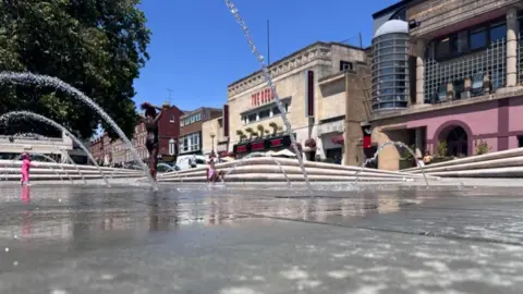A photo taken from ground level shows fountains springing from paving stones in the centre of Gloucester. Beneath cloudless skies, they spray water into the air. In the background the front of The Regal pub can be seen.