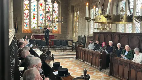 Dozens of people sat in pews at a chapel, with stained glass windows in the background.