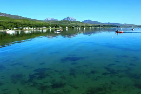 Pamela Stevenson Clear water across the sea on a sunny day, with three mountains and greenery visible behind them 