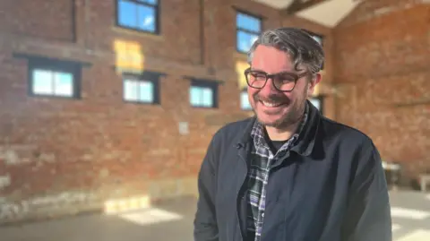 Ross Millard, smiling at the camera. He is wearing a green and purple checked shirt and a dark blue jacket. He has short wavy hair and glasses.